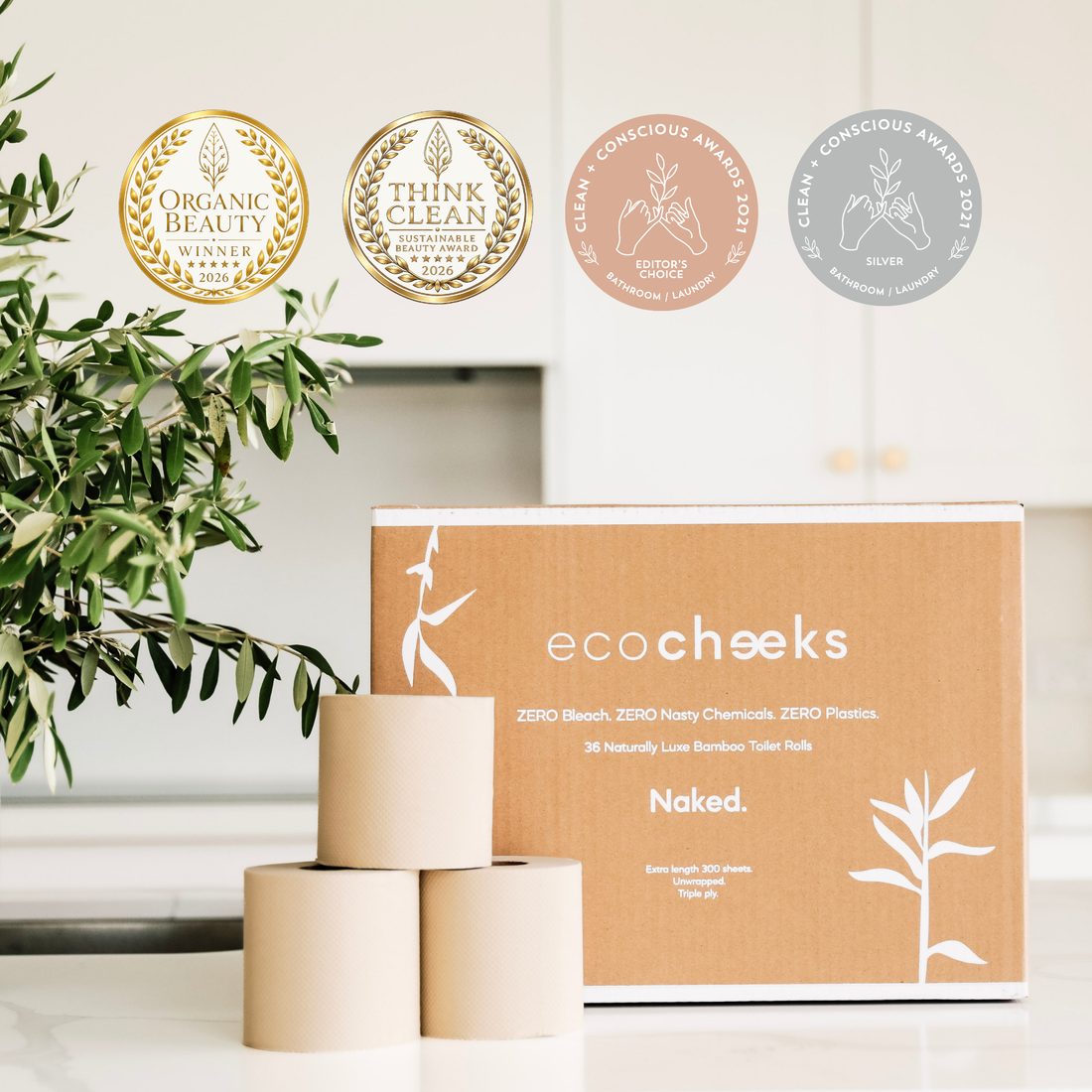 A box of ecocheeks Naked bamboo toilet paper rolls sits on a kitchen counter with three rolls stacked in front. The scene includes three award seals at the top (Organic Beauty, Think Clean, Clean Conscious Awards) and a green plant on the left.