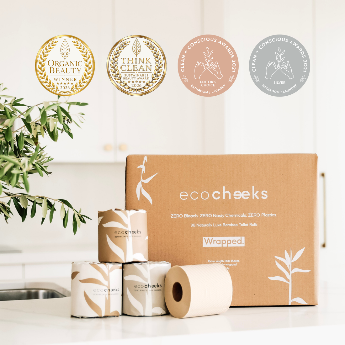 Eco Cheeks wrapped toilet paper packaging displayed on a white kitchen counter, with a large brown cardboard box, several rolls, and three award seals above. A plant is visible on the left side.