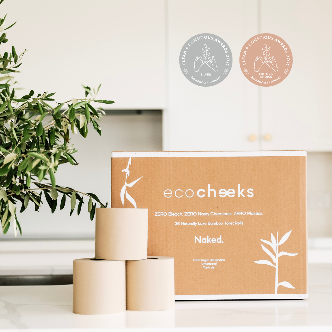 A box of eco cheeks Naked bamboo toilet rolls stands on a white countertop with three rolls stacked in front. A eco-friendly leafy green plant is on the left, and two award seals are shown in the upper right against a bright kitchen background.