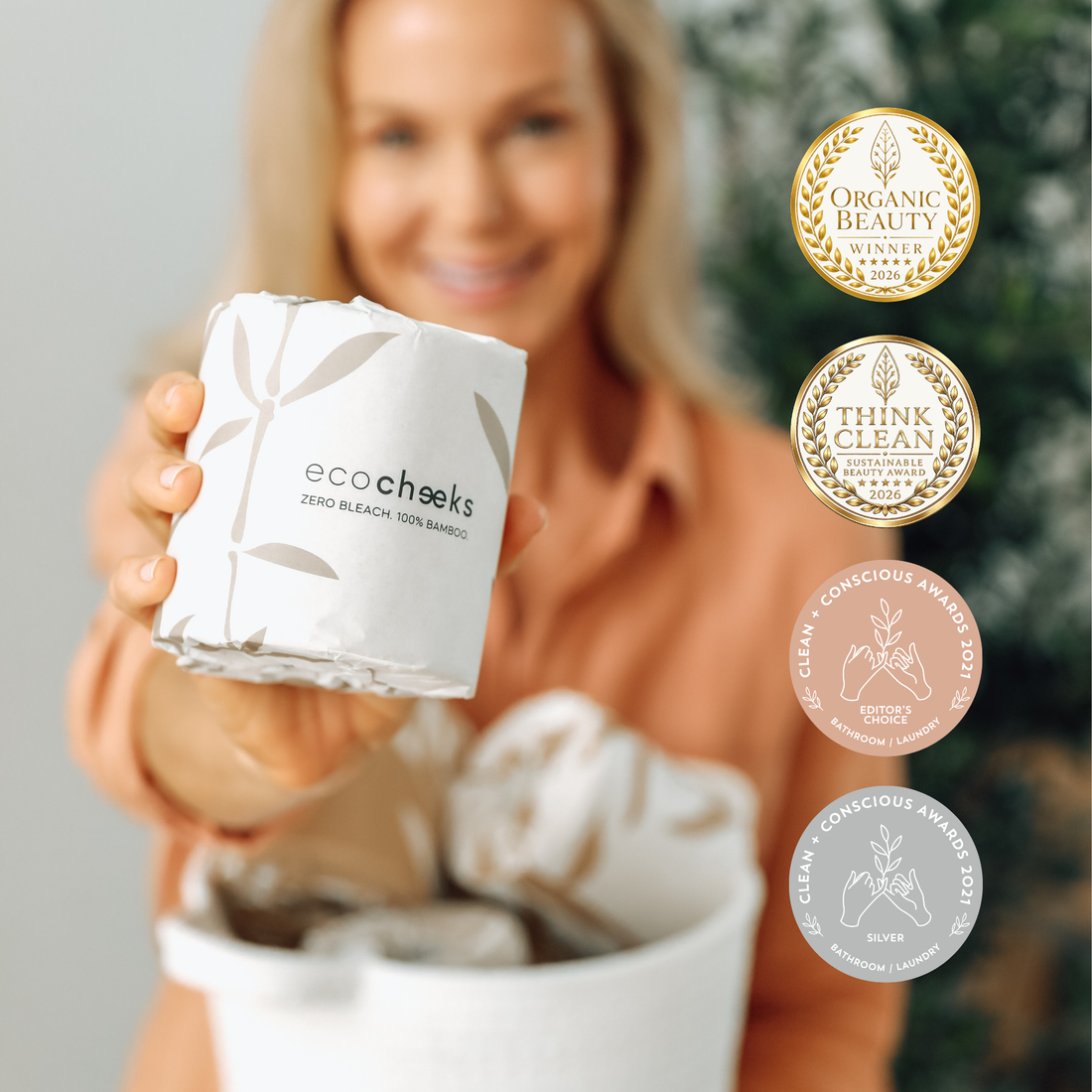 A woman holds up a white roll of eco:cheeks zero bleach 100% bamboo facial tissue. The background is heavily blurred, showing a warm indoor setting, with four circular award badges on the right side of the image.