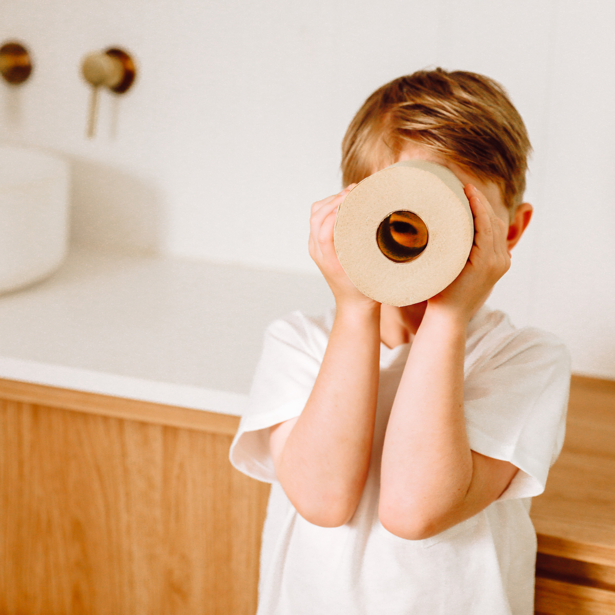 A young child with short blond hair wearing a white t-shirt holds a roll of natural brown toilet paper over their face, standing in a brightly lit bathroom with light wood cabinetry and a white sink in the background.