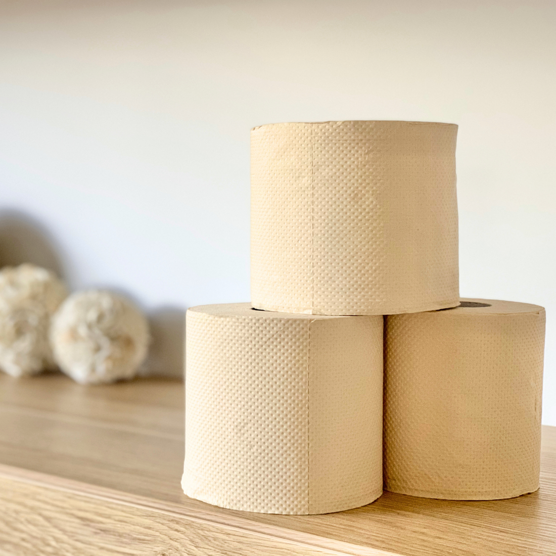 Three rolls of textured paper toilet paper stacked on a wooden surface against a soft beige background, with four circular award badges shown on the left. The badges read Organic Beauty Winner, Think Clean Winner, Clean Conscious Awards Silver, and Clean Conscious Awards Gold.